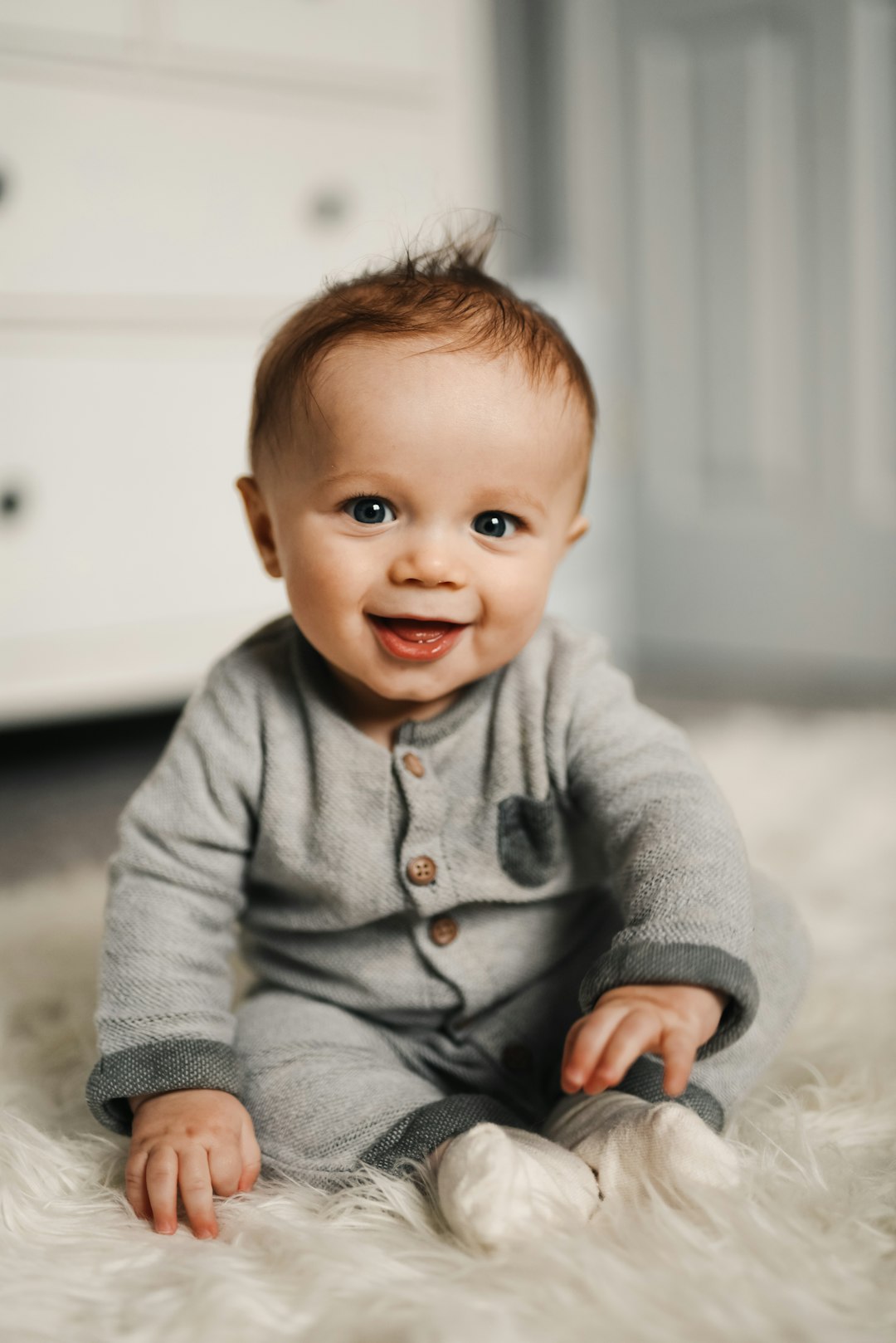 A smiling baby in a grey buttoned romper on a white faux fur rug